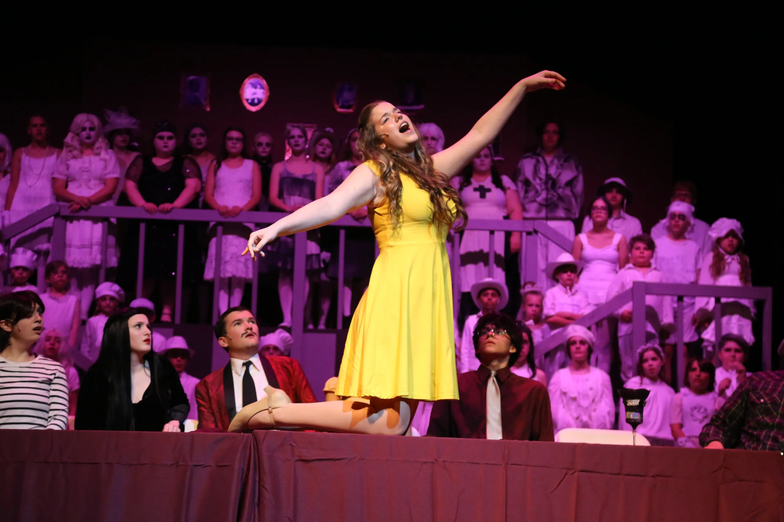 A young actress in a yellow dress performs at center stage with the full cast seated behind her