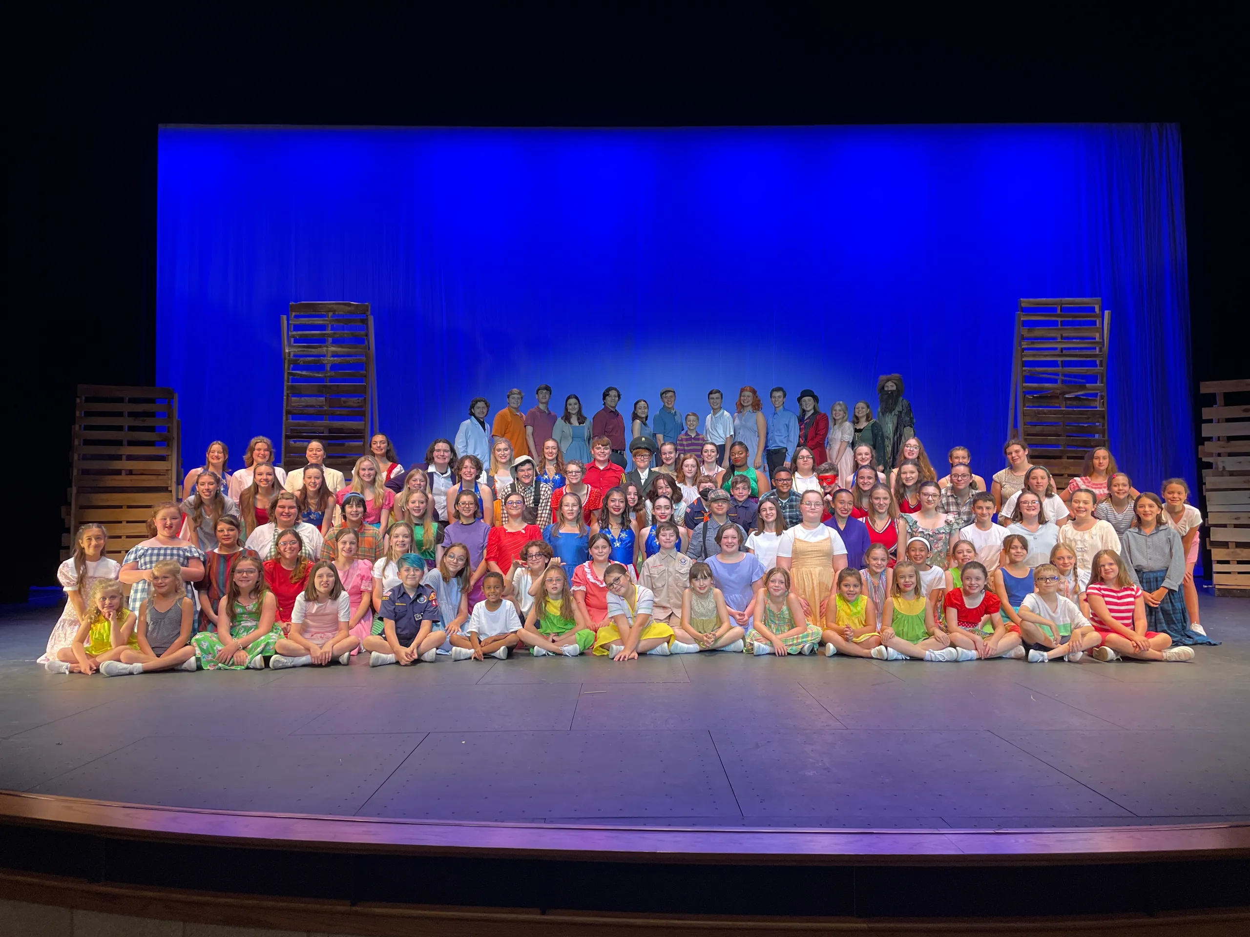 The full Big Fish cast poses together on stage in colorful costumes against a blue backdrop