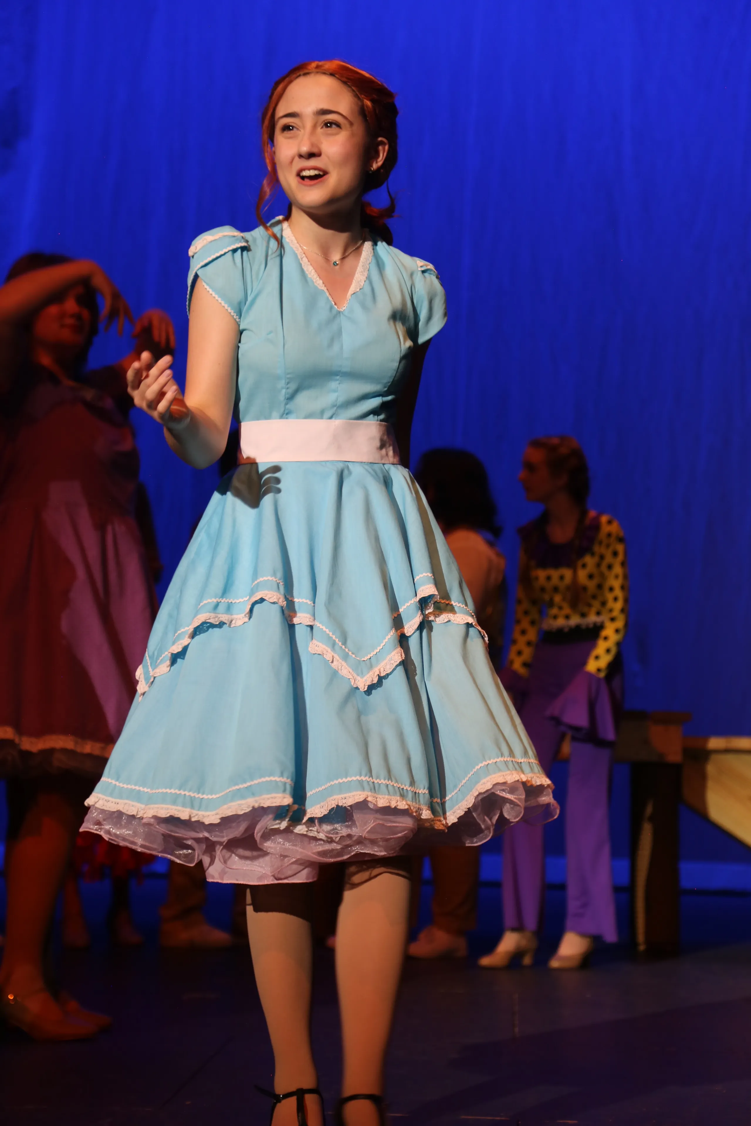A young actress in a light blue period dress sings on stage during a performance