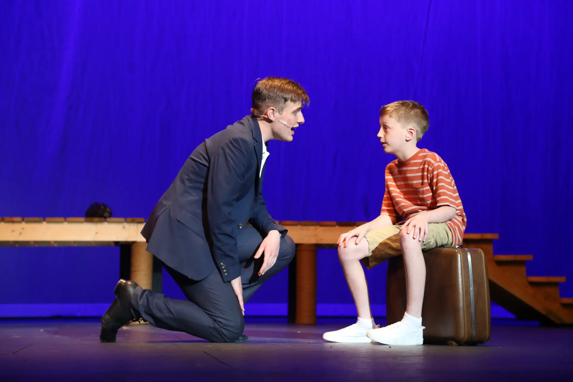 A teenage actor kneels on stage to speak with a young boy sitting on a suitcase against a blue backdrop