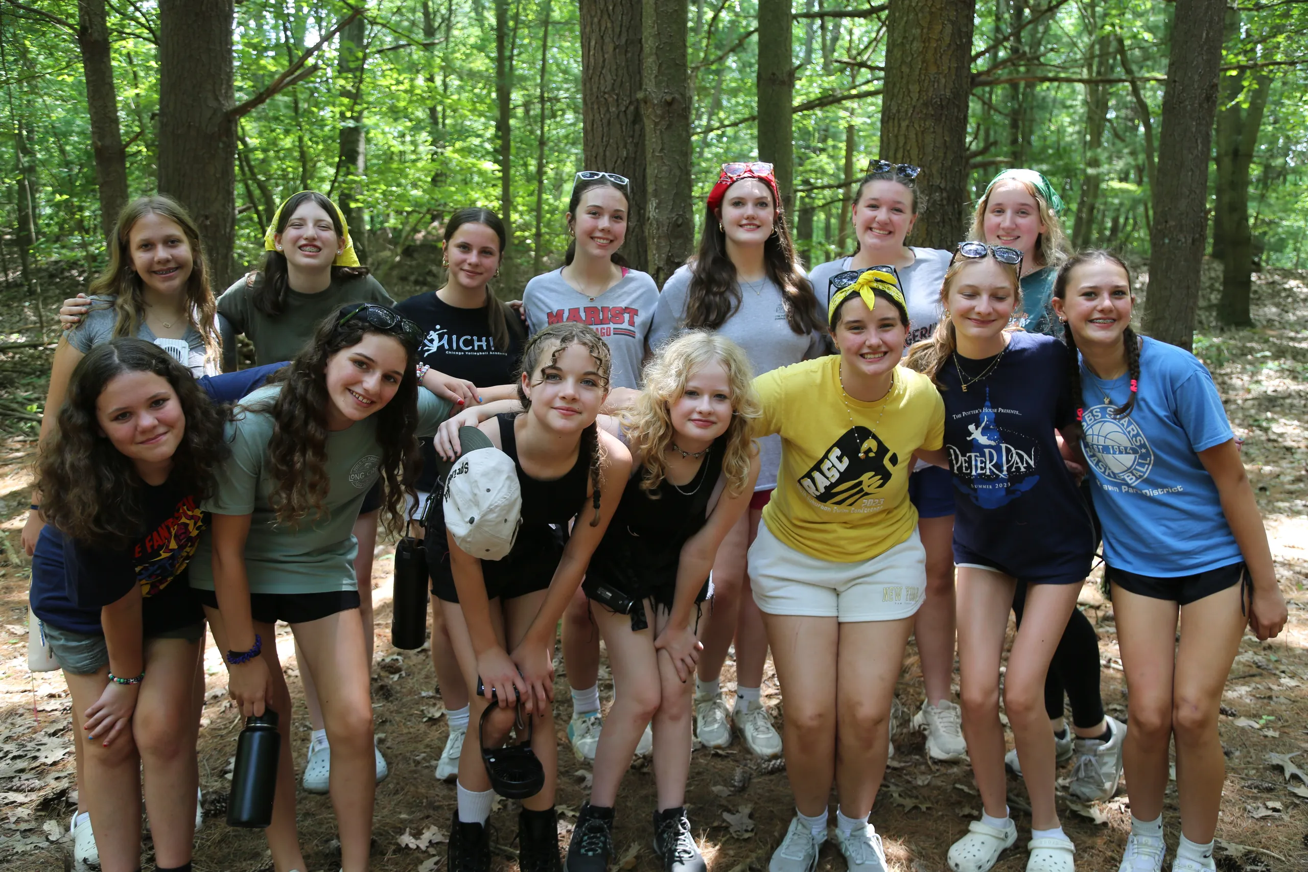 A group of teenage girls smiling together in a sunny wooded area during a camp activity
