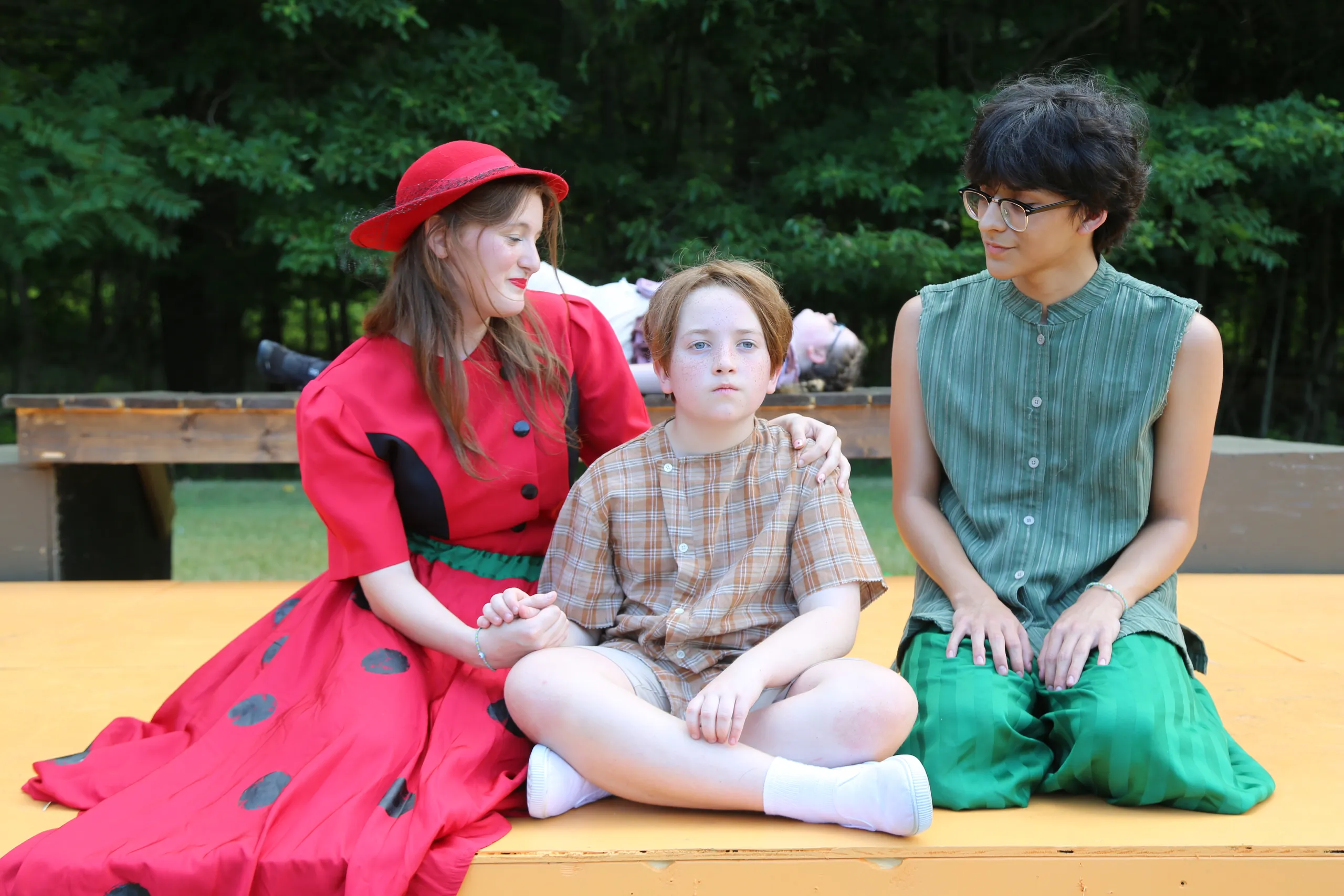 Three actors sit together on a yellow outdoor stage — two in costume flanking a young boy in the middle