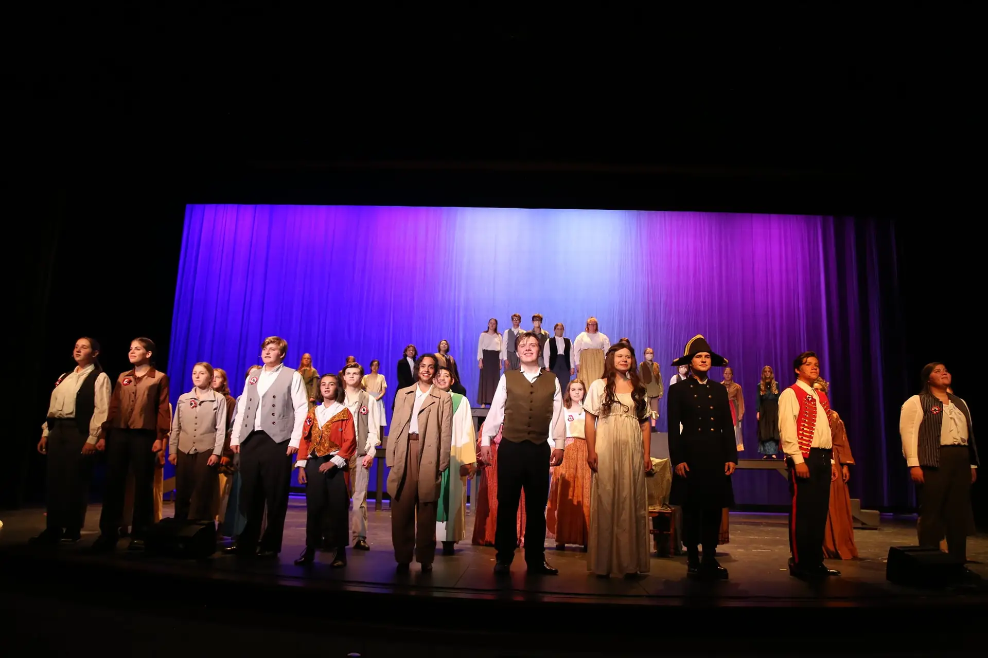 The full Les Misérables cast takes a bow on stage in period costumes under blue and purple stage lighting