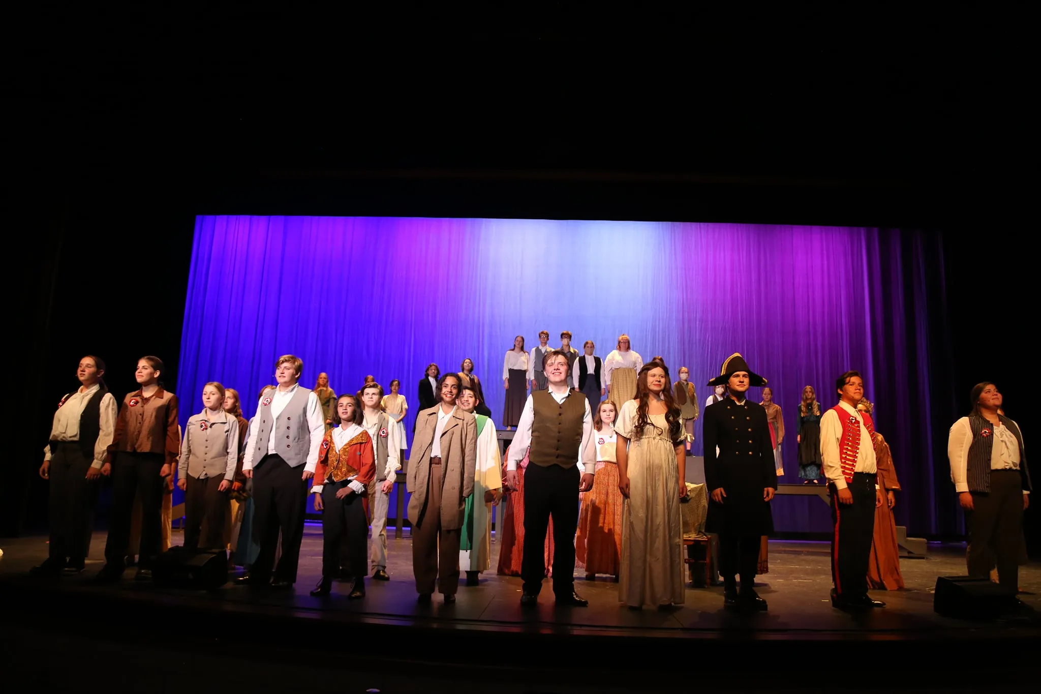The full Les Misérables cast takes a bow on stage in period costumes under blue and purple stage lighting