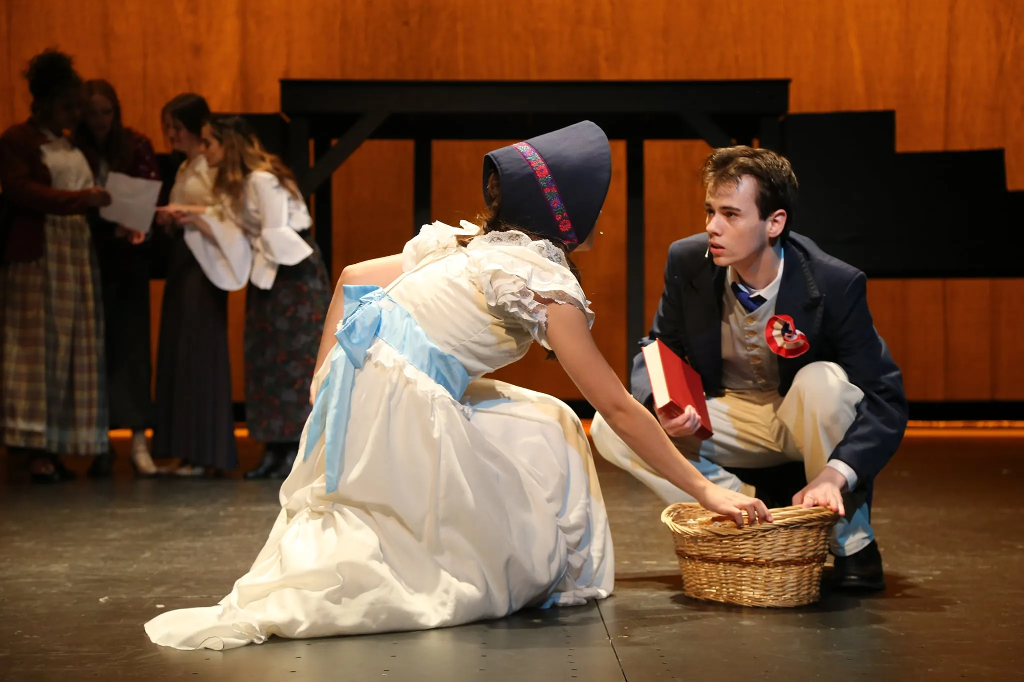 A young actress in a white dress and bonnet reaches for a basket as Marius crouches beside her during a Les Misérables scene
