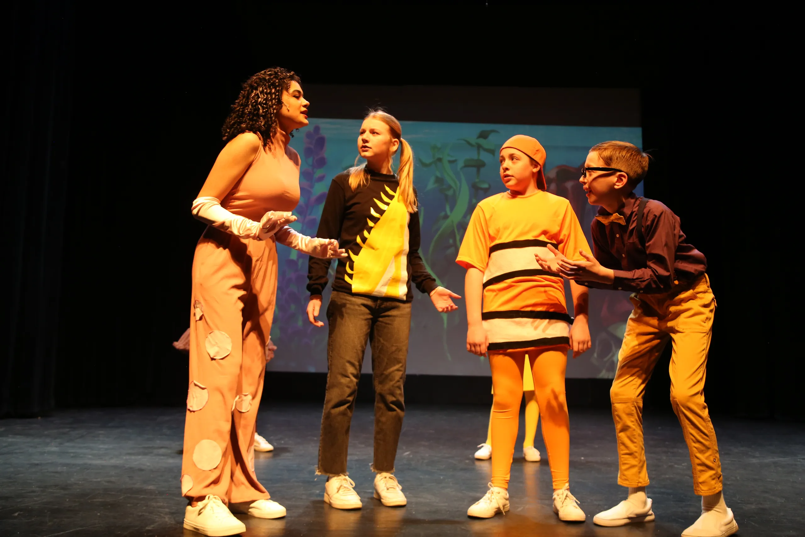 Four young actors in costume perform a scene together, with an underwater backdrop projected behind them